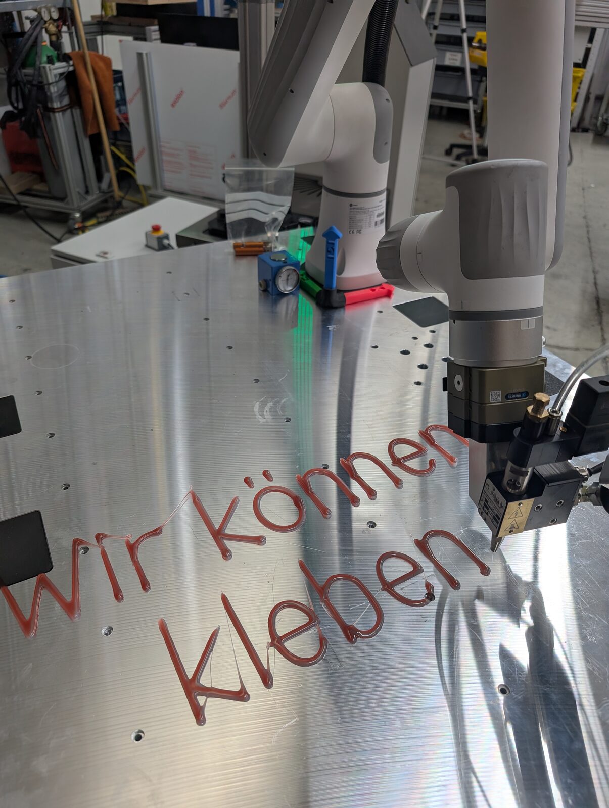 Cobot schreibt mit Dosiertechnik den Schriftzug Wir können kleben auf eine Aluminiumplatte